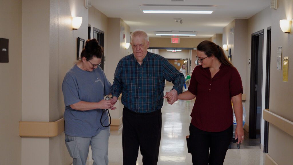A man is walking down a hallway with the assistance of two nurses who specialize in skilled nursing
