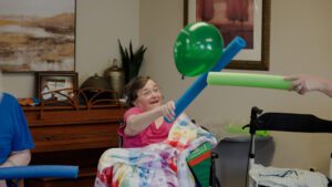 A woman plays a game with a pool noodle and balloon as an activity at a skilled nursing center