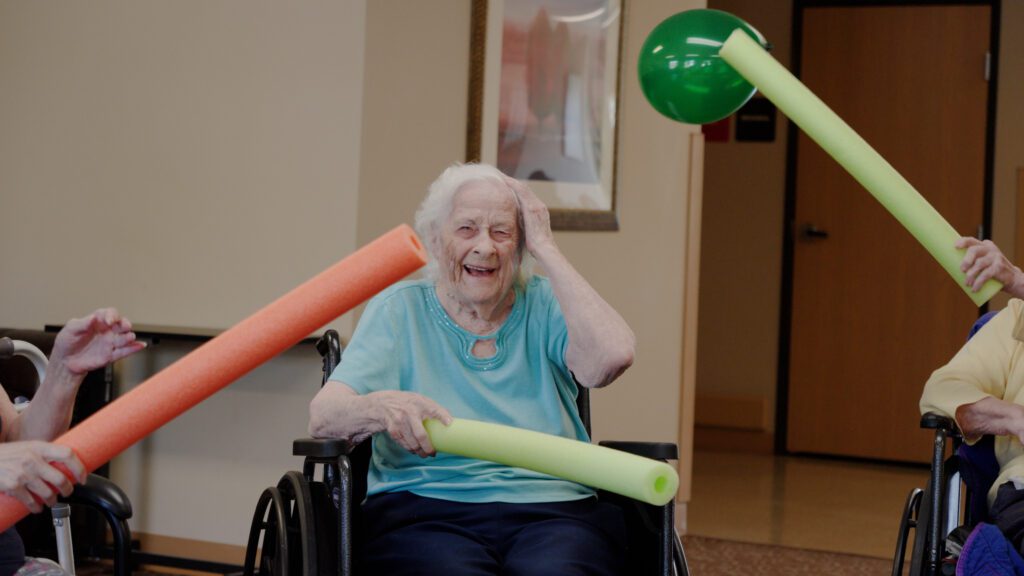A woman is smiling while playing a game with a balloon and a pool noodle at a respite care facility